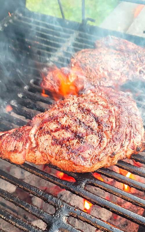 Two ribeye steaks being cooked on a cast iron grate, over charcoal.