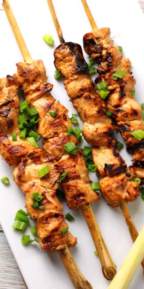 Loaded with bold flavors, these Grilled Lemongrass Chicken Skewers are a mouth-watering dish that is sure to satisfy any craving. A white serving platter is holding four skewers made from lemongrass stalks are pierced through pieces of chicken that has been grilled. Thin sliced green onions are sprinkled about as a garnish.