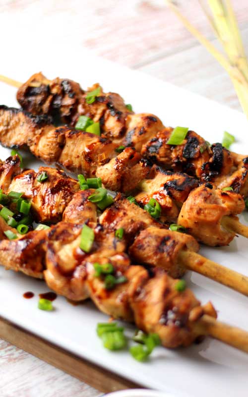 Four skewers of grilled chicken are on a white serving platter. The chicken has been garnished with thin sliced green onions.