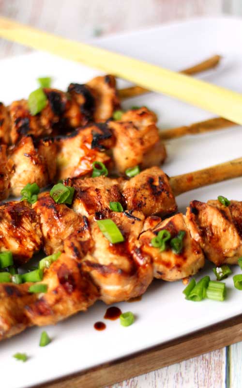 Four skewers of grilled chicken are resting on a white platter. Green onions are used as a garnish on the chicken.