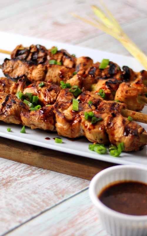 Four skewers of grilled chicken, garnished with green onions, are arranged on a white serving platter. A small white dish is filled with a dipping sauce, and arranged in the bottom-right corner of the image.