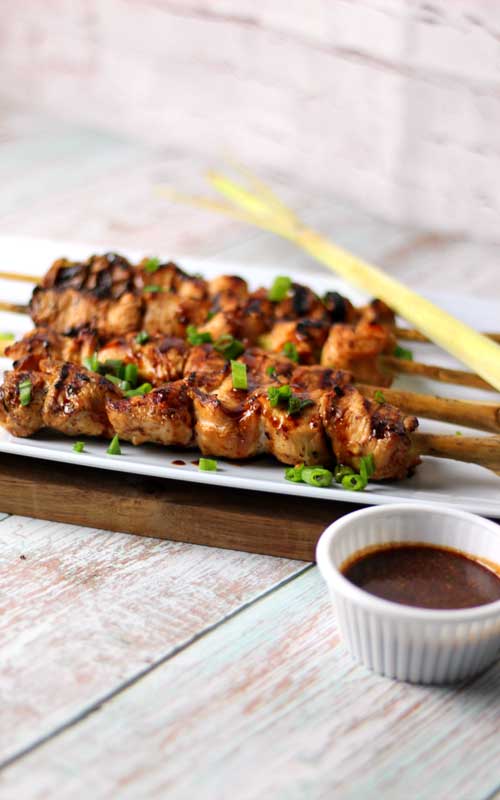 A white serving platter rests upon a thin piece of wood. On the platter are four grilled lemongrass chicken skewers. The chicken has been garnished with green onion. To the front-right of the platter is a small cup of a brown dipping sauce.