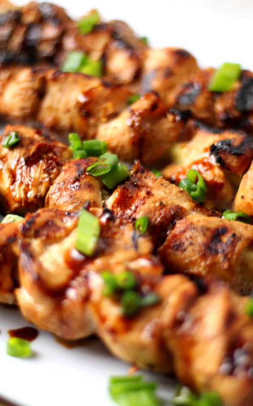 A close up view of chicken that has been cubed then grilled. The chicken is garnished with green onion, and resting on a white dish.