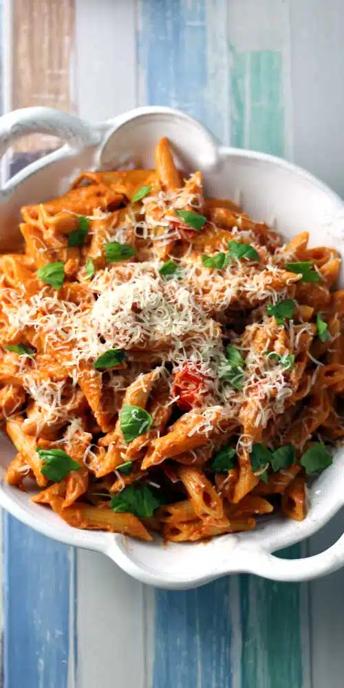 A top down view of a white bowl filled with penne pasta in a tomato sauce. The background is wood painted in green, blue, and white stripes.