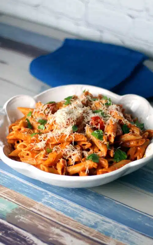 Penne pasta in a tomato sauce in a white serving dish. The dish is sitting on a multi-colored piece of wood, with a blue towel in the background.