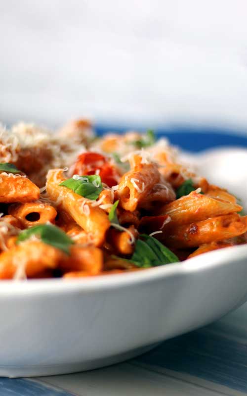 A close up view of a white serving dish. In the dish is a tomato covered penne pasta with pieces of basil.