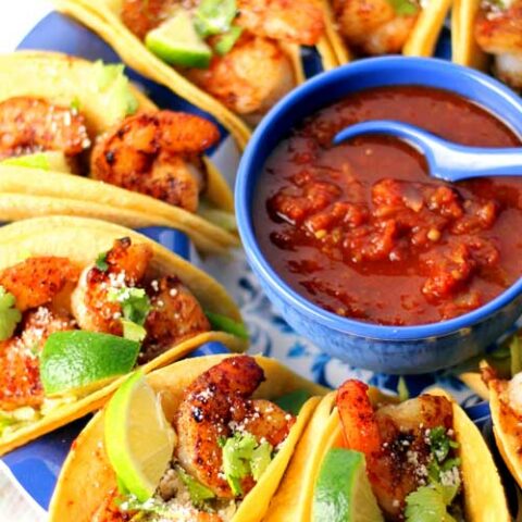 Top down view of shrimp tacos on a round plate. In the center of the plate is a blue bowl filled with salsa.