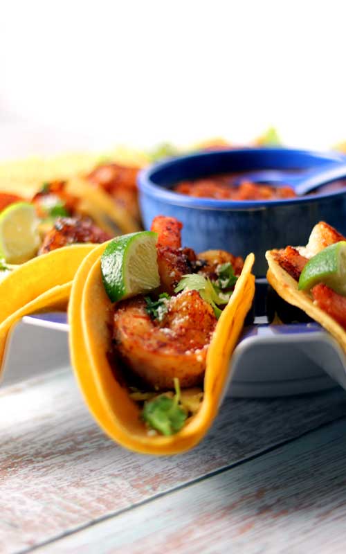 View of the end of a shrimp street taco. The taco is standing up on a plate. In the background is a blue bowl filled with salsa.