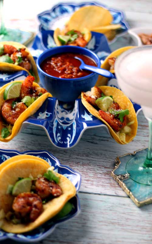 Shrimp tacos on a round platter. In the center of the platter is a blue bowl filled with salsa. On the right edge of the photo is a frown magarita in a glass.