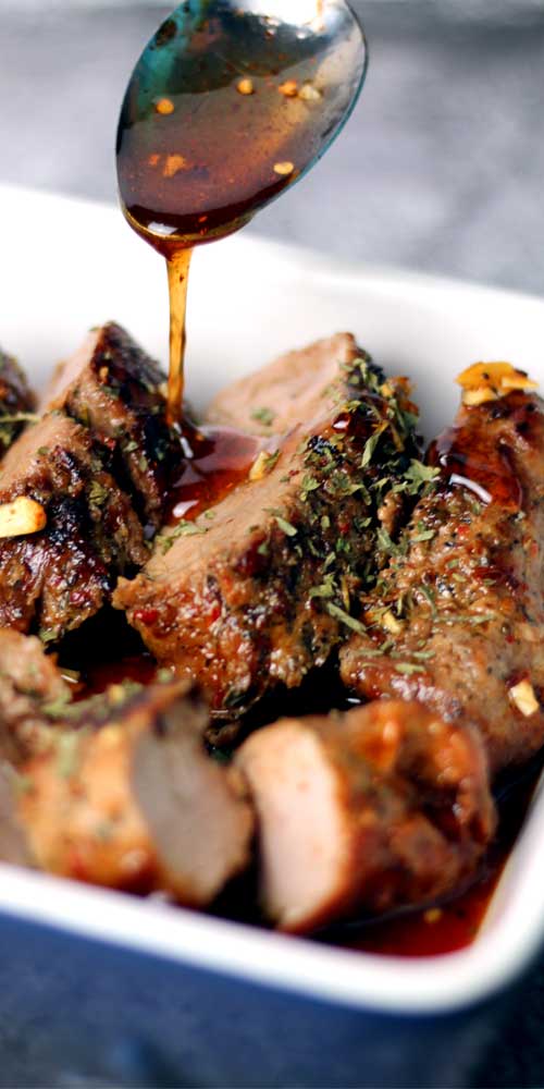 A metal spoon pouring hot honey onto a cooked and sliced pork tenderloin. The pork tenderloin is in a white dish.