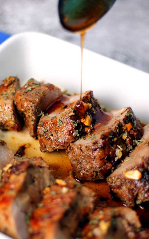 Hot honey being drizzled on top of a cooked, sliced pork tenderloin that is in a white dish.