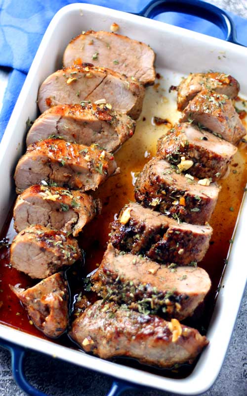 An overhead image of a sliced and cooked pork tenderloin in a white baking dish. The tenderloins natural juices ar pooling up in 2 corners of the dish.