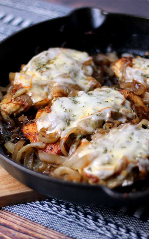 A cast iron skillet containing caramelized onions and cooked chicken breasts. The chicken is covered in melted Muenster cheese.