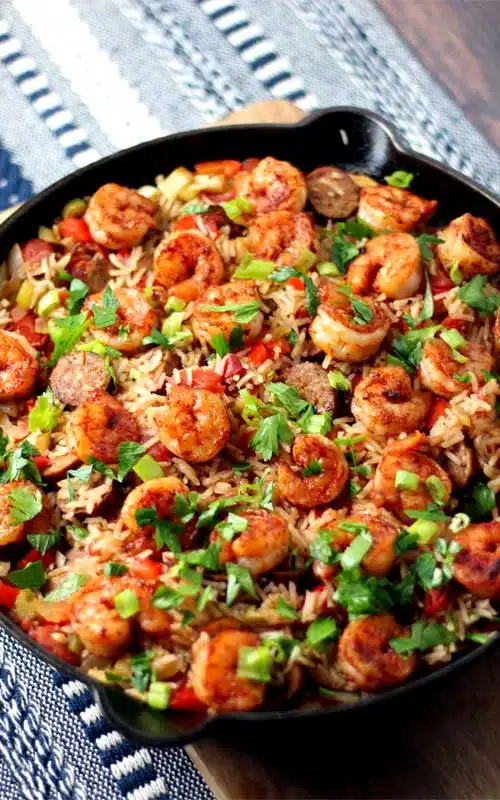 A cast iron skillet filled with cooked rice, shrimp, and sausage. The food is garnished with parsley that has been coarsely torn.