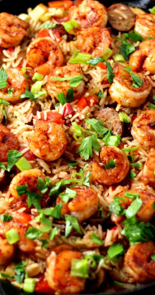 Shrimp, sausage, and cooked rice garnished with parsley. Red and green bell peppers can also be seen spread throughout the dish.