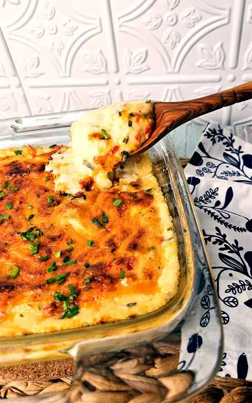 A wooden spoon scooping mashed potatoes from a clear baking dish. The potatoes are topped with melted cheese and chives.