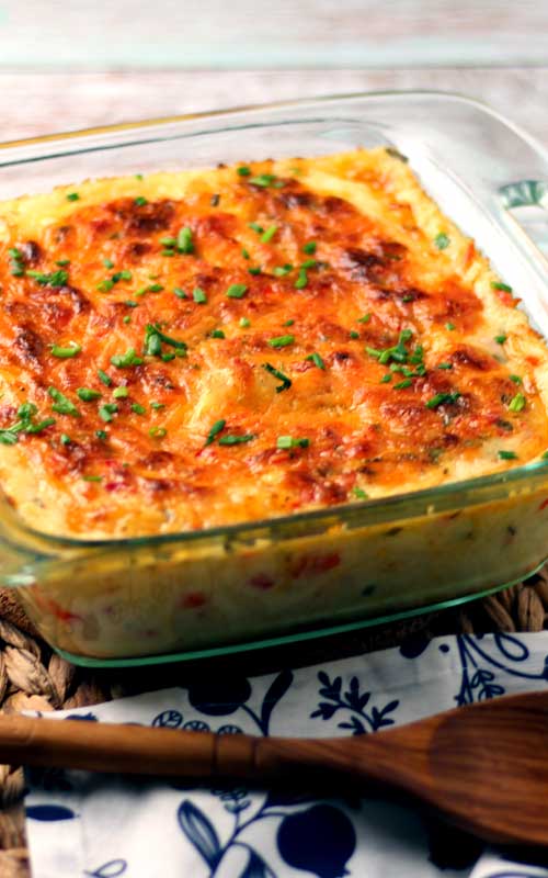 Melted and browned cheese along with chives top baked mashed potatoes that are in a clear baking dish.