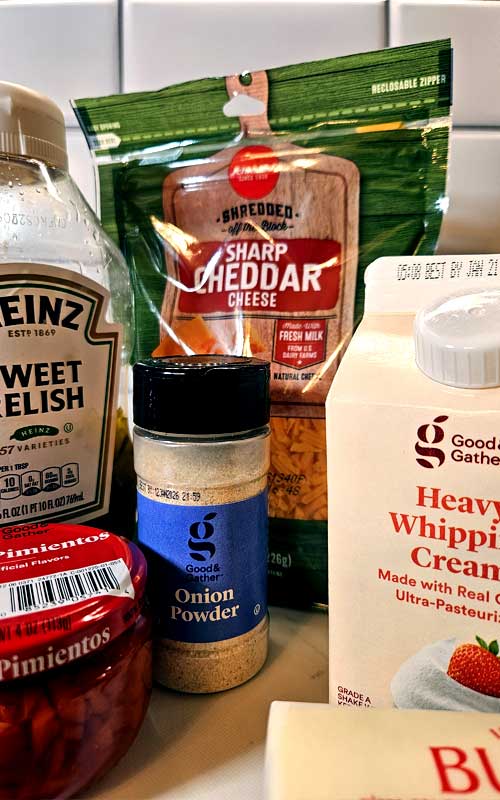 Packaged ingredients used to make Pimento Cheese Mashed Potatoes. Items included are: pickle relish, a bag of shredded cheddar cheese, a jar of pimentos, a shaker of onion powder, a carton of heavy whipping cream, and a stick of butter.