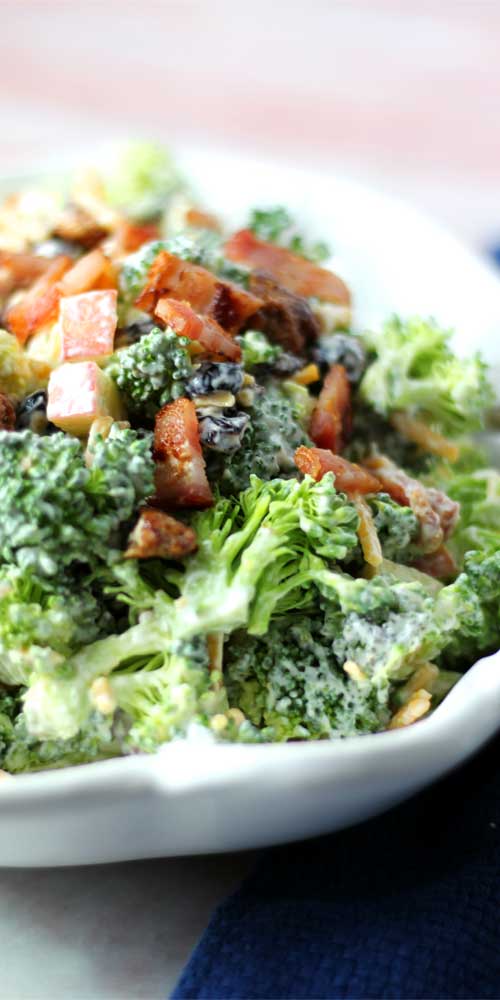Near over head view of a white bowl with a salad prepared using broccoli, apples, cheddar cheese, and bacon.