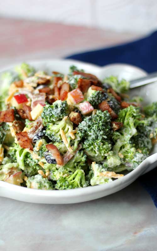 Broccoli, bacon, apples, and cheddar cheese mixed together into a salad. Everything is arranged in a white bowl. A metal spoon can be seen coming out of the bowl.