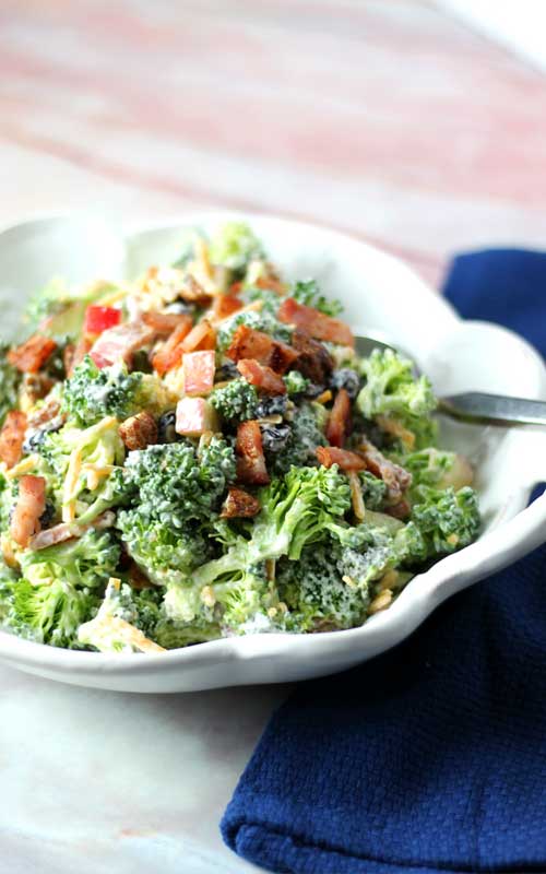 A white bowl containing a prepared broccoli salad. The salad is made up of broccoli, bacon, diced apples, and cheddar cheese.