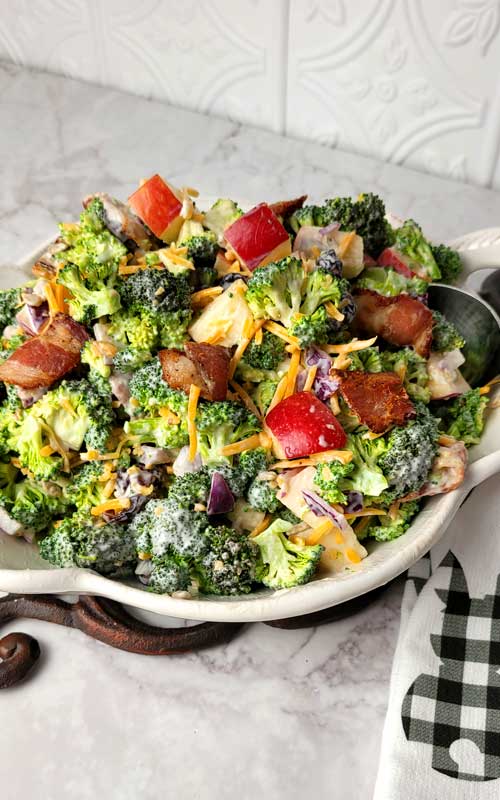 Broccoli, apples, bacon, and cheddar cheese mixed together into a salad. Everything is arranged in a white bowl.