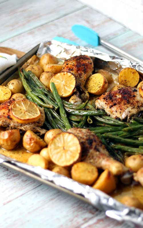 A sheet pan lined with aluminum foil. On the foil is cooked chicken pieces, lemons, green beans, and halved creamer potatoes.