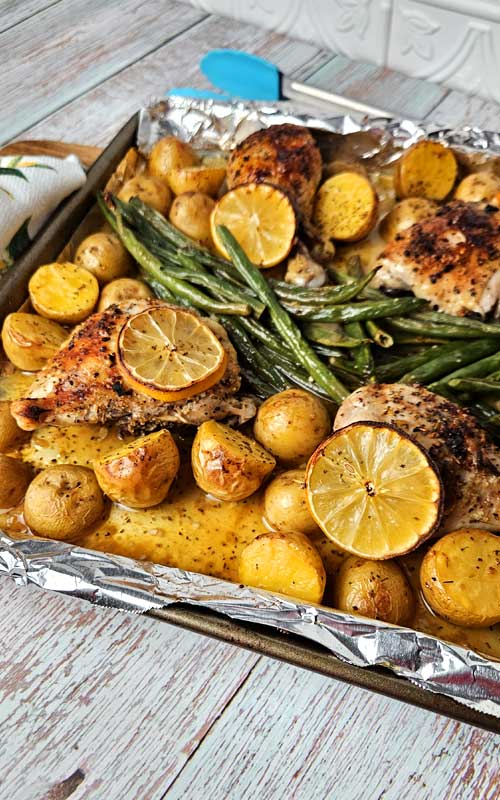 A sheet pan lined with aluminum foil. On the foil is cooked chicken pieces, lemons, green beans, and halved creamer potatoes.