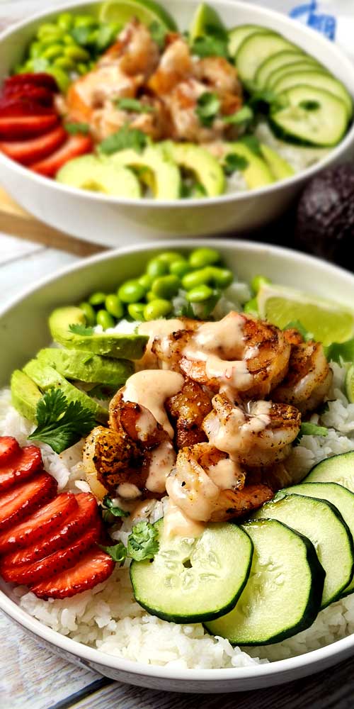 Two white bowls filled with prepared rice. The rice is topped with sliced strawberries, sliced avocado, and edamame. In the center of each bowl is a mound of shrimp covered in a peach colored sauce.