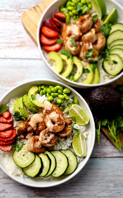 Two white bowls filled with shrimp, white rice, cucumber, avocado, strawberries, and edamame.
