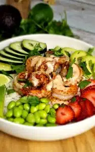 A wide, white bowl filled with shrimp, rice, strawberries, sliced cucumber, edamame, and sliced avocado.